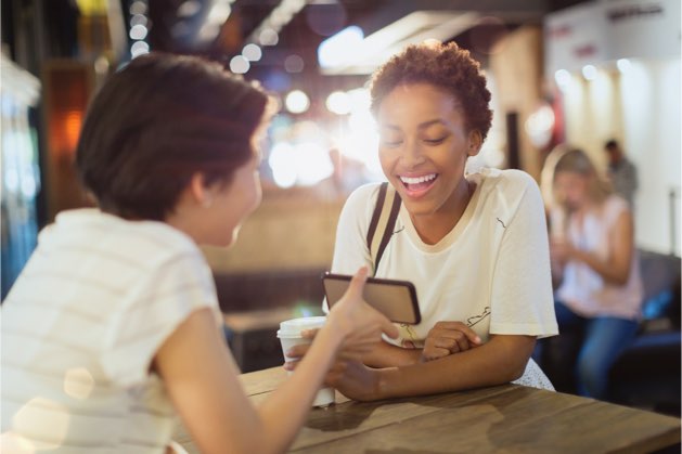 A young women holding out her phone, showing it to her smiling friend.