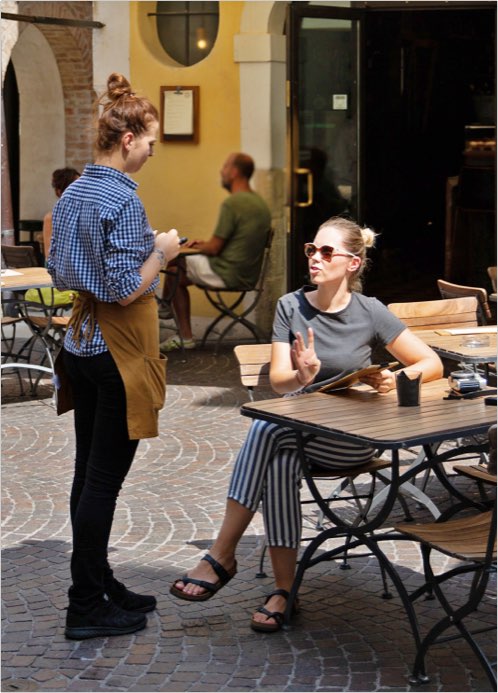 A young woman sits at a cafe table, speaking to a waitress.